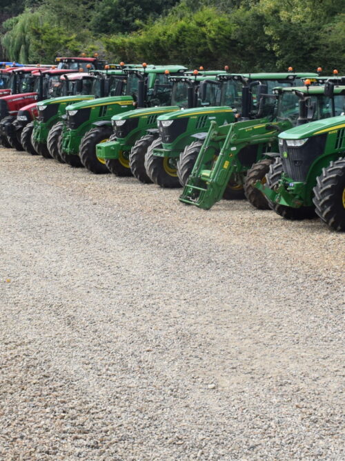 A row of various tractors neatly lined up on a gravel lot. Most tractors are green with yellow wheels, indicating they are John Deere models. There are also red, blue, and yellow tractors further down the line, alongside other agricultural equipment.