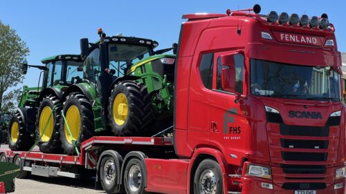 2 Green and yellow John Deere tractors sitting on the bed of a red low loader lorry
